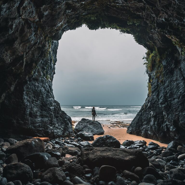 Blick aus einer Steinhöle hinaus. Der Ausgang ist riesig und vor der Höle ist das Strand und Meer zu sehen